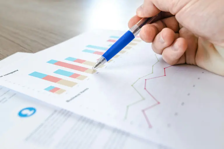 A hand holding a blue pen points to a colorful bar graph on a sheet of paper. The graph includes blue, red, and beige bars, with another line graph in green and red visible in the background. The document likely relates to business metrics, including a click-through rate analysis.