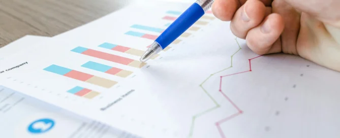 A hand holding a blue pen points to a colorful bar graph on a sheet of paper. The graph includes blue, red, and beige bars, with another line graph in green and red visible in the background. The document likely relates to business metrics, including a click-through rate analysis.