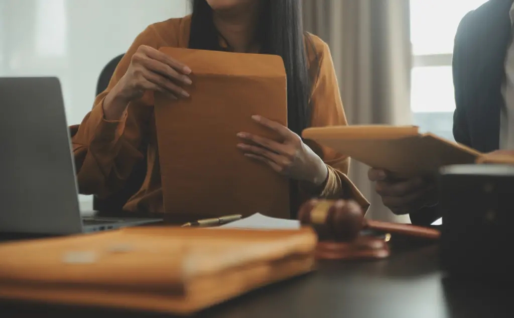 Two individuals handling large brown envelopes at a desk with a laptop and a judge's gavel, representing legal counsel and documentation.