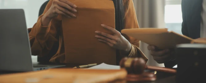 Two individuals handling large brown envelopes at a desk with a laptop and a judge's gavel, representing legal counsel and documentation.