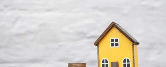 Miniature yellow house next to stacks of coins, symbolising property value and costs of selling a home.