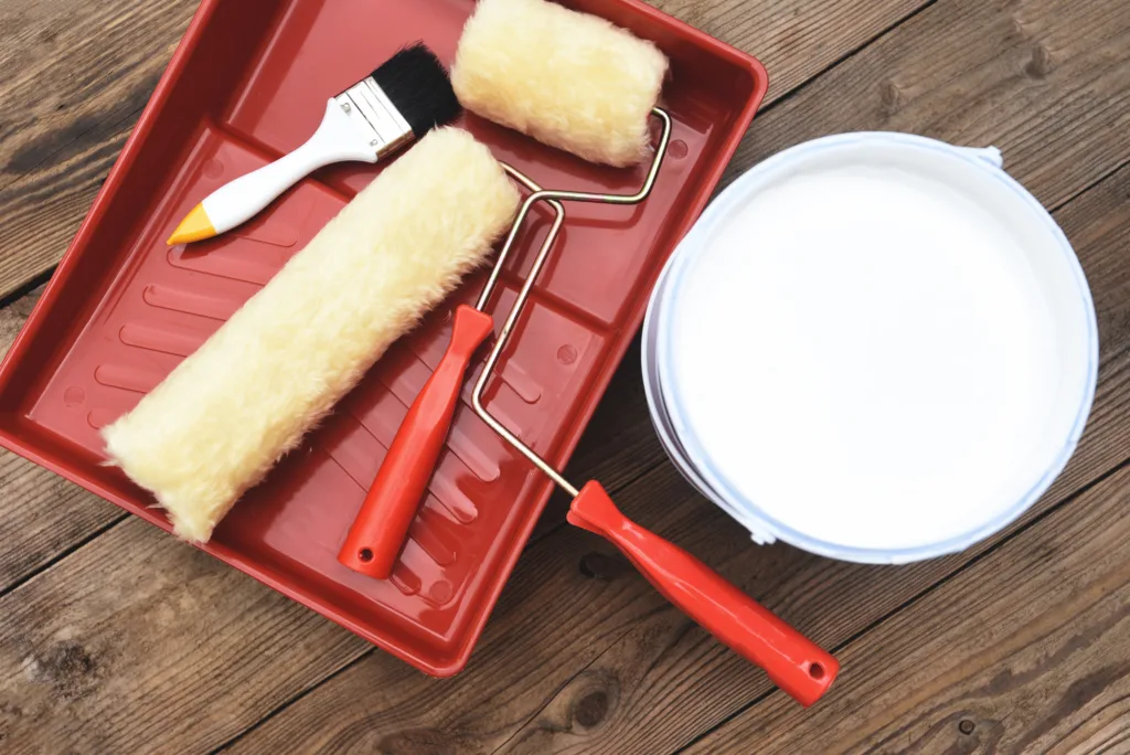 Painting tools including a roller, brush, tray, and a can of white paint on a wooden floor.