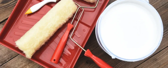 Painting tools including a roller, brush, tray, and a can of white paint on a wooden floor.