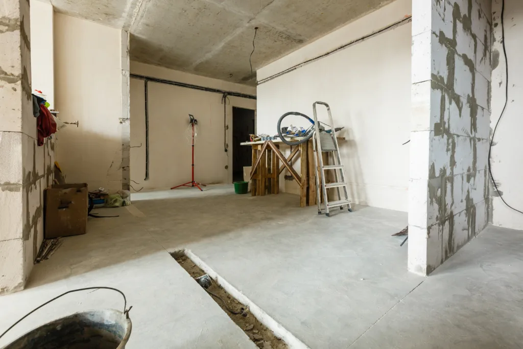 The image shows a room under construction or renovation, with unfinished walls and exposed electrical wiring. There's a ladder leaning against the wall, a workbench with tools, and construction materials scattered around the area.