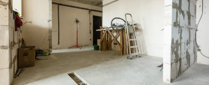 The image shows a room under construction or renovation, with unfinished walls and exposed electrical wiring. There's a ladder leaning against the wall, a workbench with tools, and construction materials scattered around the area.