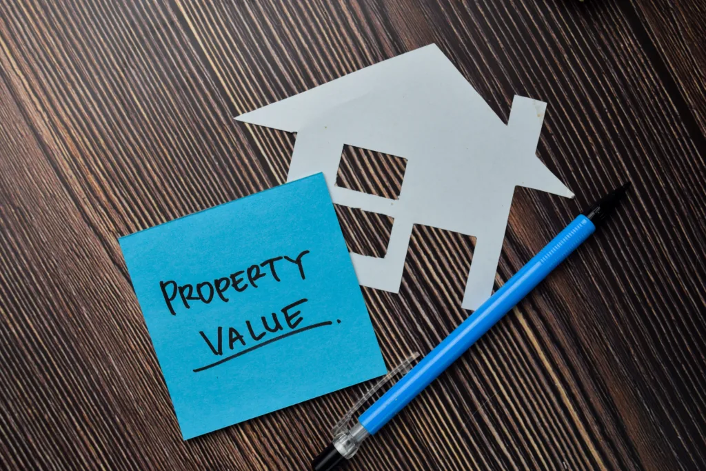 Sticky note with 'Property Value' written on it next to a paper cutout of a house and a blue pen on a wooden surface.