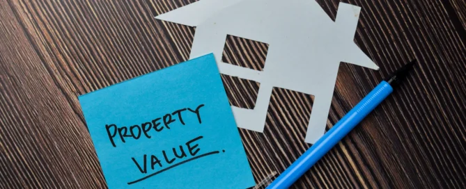 Sticky note with 'Property Value' written on it next to a paper cutout of a house and a blue pen on a wooden surface.