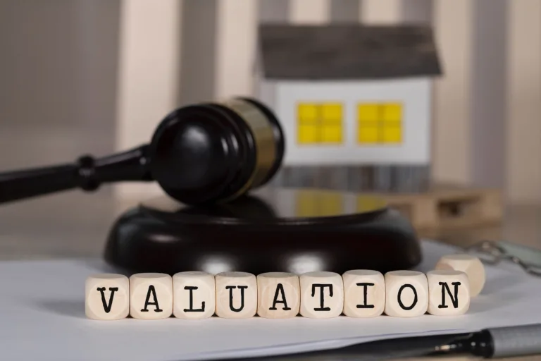 The image shows wooden blocks spelling out the word "VALUATION" placed in front of a small house model and a gavel, symbolising property valuation and legal considerations.