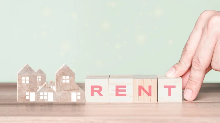 Hand placing wooden blocks spelling 'RENT' next to small wooden house models