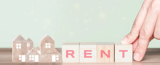 Hand placing wooden blocks spelling 'RENT' next to small wooden house models