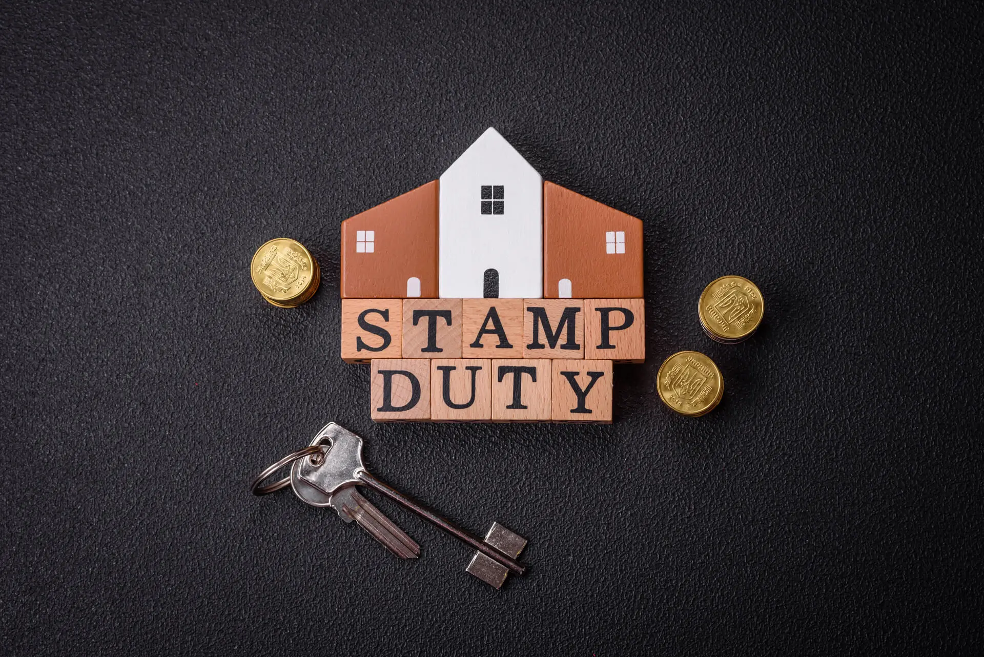 Stamp duty concept with a house model, wooden letter blocks spelling 'STAMP DUTY,' British pound coins, and house keys on a dark background.