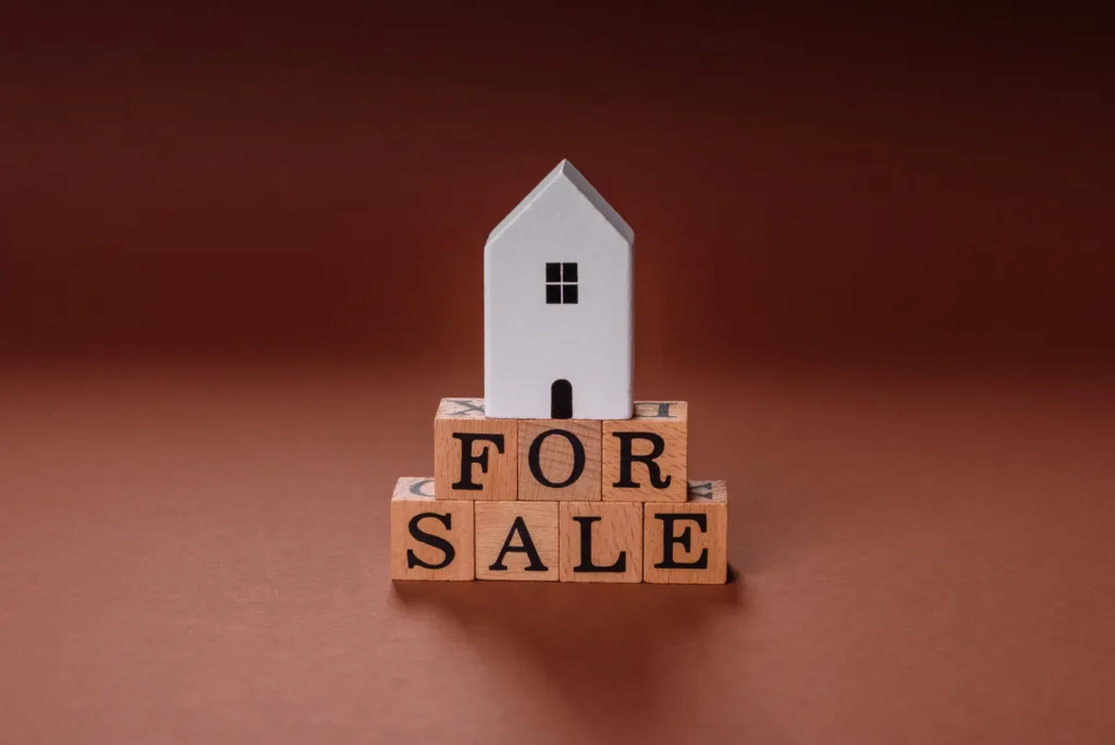 White model house sitting on wooden letter blocks spelling "FOR SALE" against warm brown background