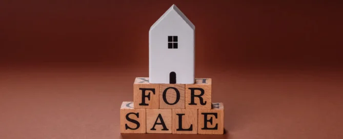 White model house sitting on wooden letter blocks spelling "FOR SALE" against warm brown background