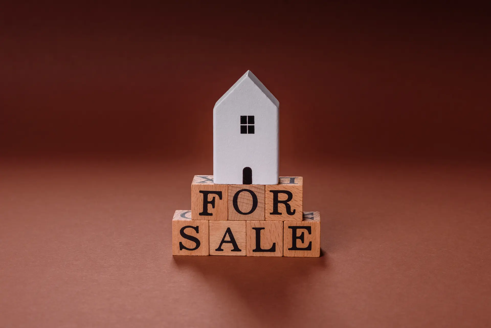 White model house sitting on wooden letter blocks spelling "FOR SALE" against warm brown background