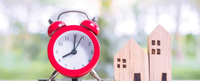 Red alarm clock next to wooden model houses on stacked coins representing property timing and investment decisions