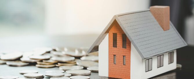 Model house placed beside scattered coins on a table, representing home costs and real estate finances.