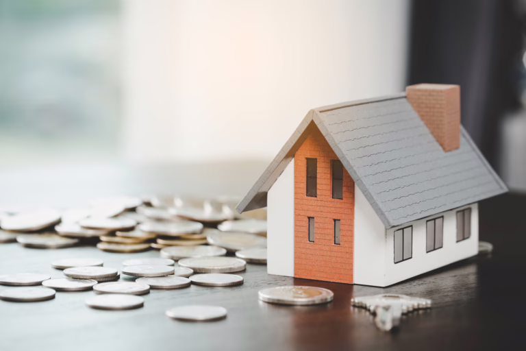 Model house placed beside scattered coins on a table, representing home costs and real estate finances.
