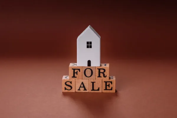 White model house sitting on wooden letter blocks spelling "FOR SALE" against warm brown background