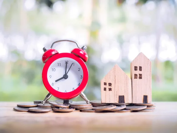 Red alarm clock next to wooden model houses on stacked coins representing property timing and investment decisions